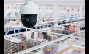A white and black dome-shaped CCTV camera hangs from the ceiling, overlooking a blurred, expansive retail store or warehouse interior with aisles and products.