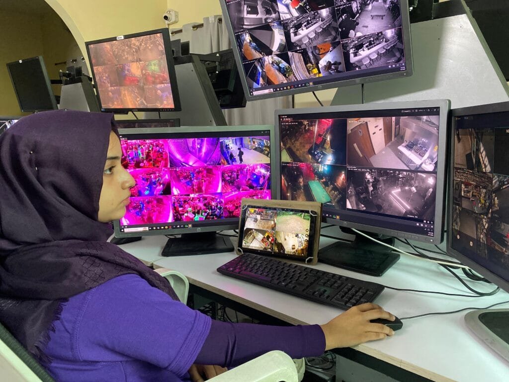 Female security operator in a hijab monitoring a multi-screen CCTV Monitoring station, using a mouse and keyboard to manage footage for timely Workplace Incident Report generation.