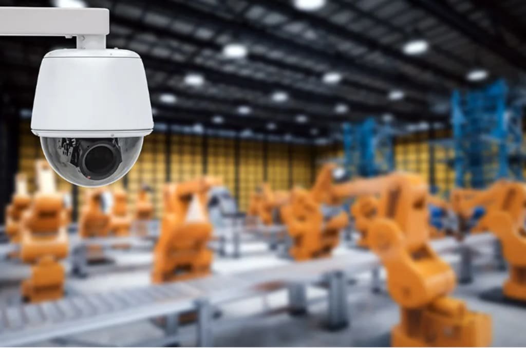 A white, spherical CCTV dome camera is mounted on a white arm in the upper left foreground, sharply in focus. Its dark lens is visible, facing towards the right. The background is a blurred yet discernible image of a large industrial facility or warehouse. Rows of orange robotic arms or machinery are visible on a conveyor system, stretching into the distance under overhead lighting. The overall impression highlights the integration of advanced CCTV monitoring and remote monitoring systems for robust intruder prevention, ensuring 24/7 monitoring and enhanced business security in an industrial or warehouse setting.