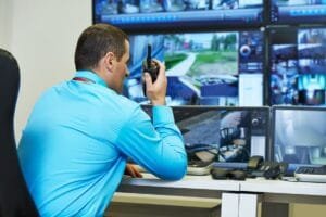 Security operator in a control room monitoring live CCTV feeds on multiple screens while communicating via radio. A professional security operator actively monitors multiple camera feeds using a walkie-talkie, demonstrating the effectiveness of 24/7 live CCTV monitoring.