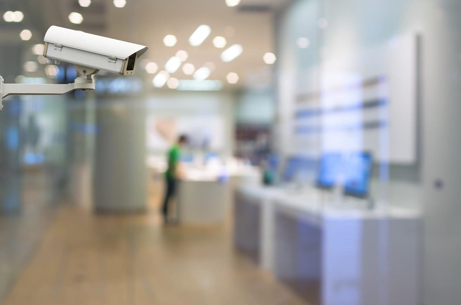 A white outdoor bullet-style CCTV camera is mounted on a wall, monitoring the entrance of a modern retail store with a customer visible inside. The store has bright, blurred lights and a glass front.