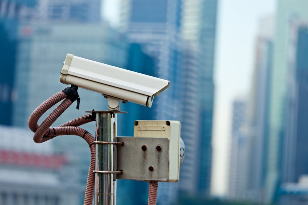 Outdoor bullet-style security camera mounted on a pole with a blurred cityscape background, symbolizing Remote CCTV Monitoring Services for businesses across the USA.