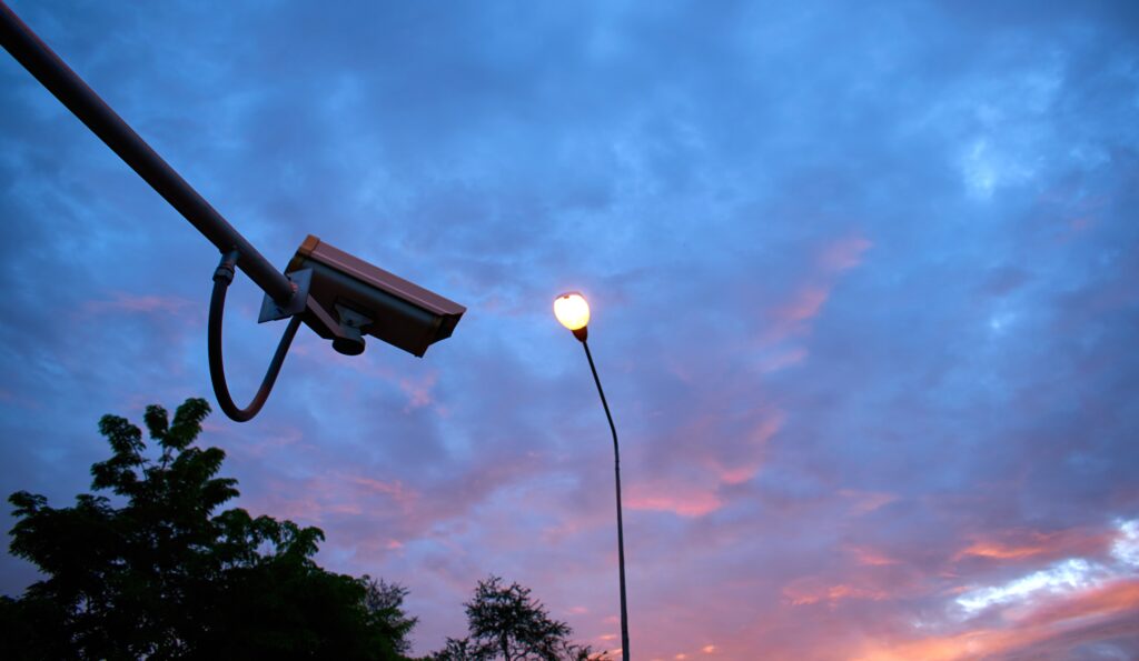 Security camera mounted on a pole next to a streetlight at dusk, representing remote monitoring services and 24/7 CCTV monitoring services.


