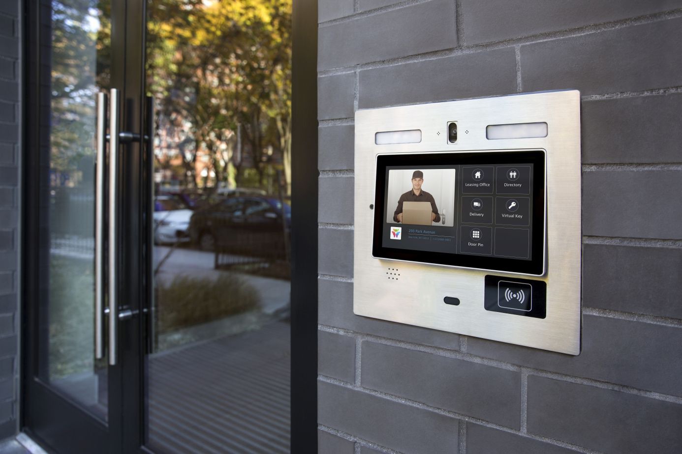 A close-up of a brushed metal intercom and display panel mounted on a brick wall next to a glass door. The screen shows a live video feed of a man holding a package, demonstrating a key function of a Virtual Doorman.