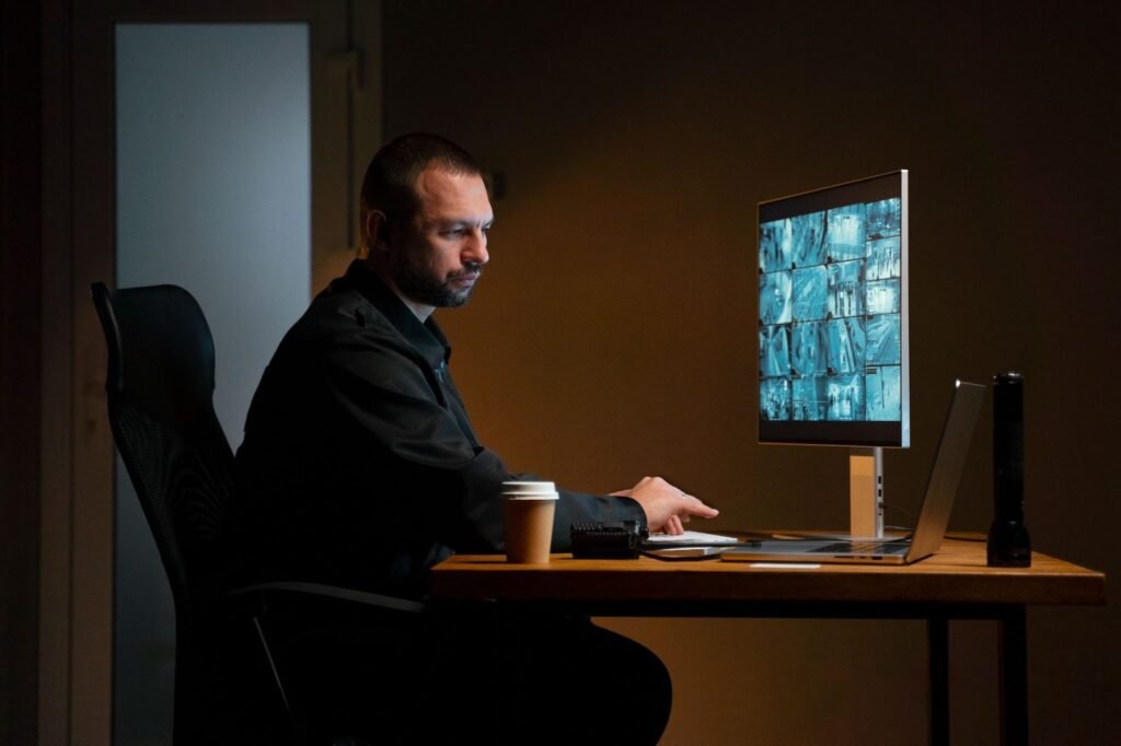A security guard or operator sitting in a dark room, intently focused on a computer screen displaying multiple split-screen CCTV monitoring feeds.