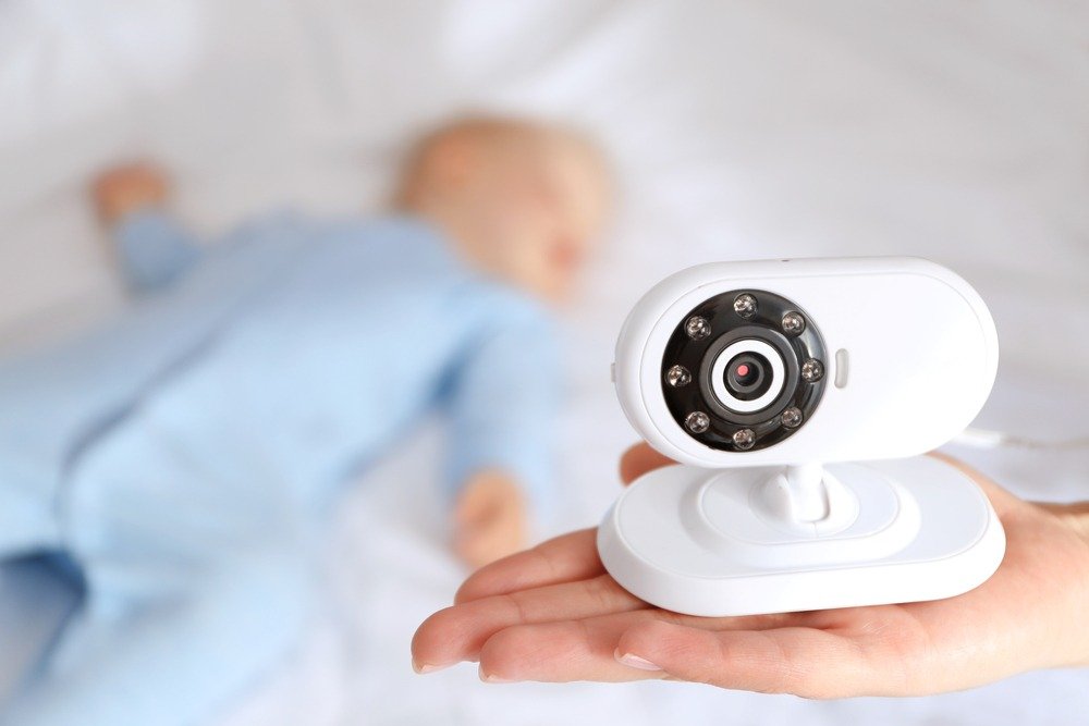 Close-up of a small white nanny camera in a person’s palm, with a sleeping baby softly blurred in the background, symbolizing safe, discreet home monitoring.