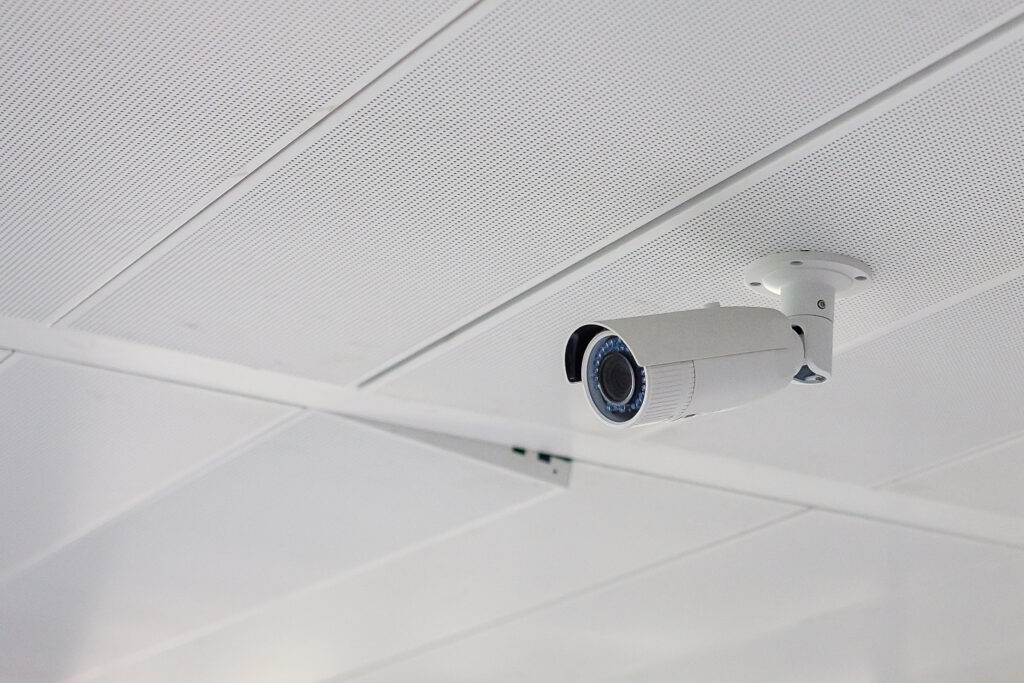 A white bullet-style security camera mounted on a suspended acoustic tile ceiling in a bright, modern interior. The installation reflects the precision of top surveillance providers.
