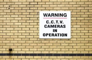 White warning sign on a brick wall reading "C.C.T.V. Cameras in Operation," confirming CCTV Surveillance.