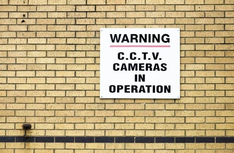 White warning sign on a brick wall reading "C.C.T.V. Cameras in Operation," confirming CCTV Surveillance.