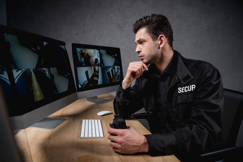 Male security officer in uniform observing multi-view monitors while holding a radio, representing Professional Security Monitoring.