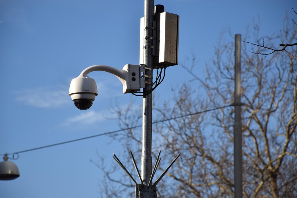 A white, adjustable outdoor CCTV camera mounted on a tall metal pole alongside a utility box, set against a blue sky, symbolizing the equipment used by a CCTV monitoring company.