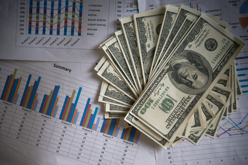 Fanned out $100 US bills placed on top of several sheets of paper displaying colorful business charts, graphs, and sales summaries, representing the CCTV monitoring cost and financial investment in security.