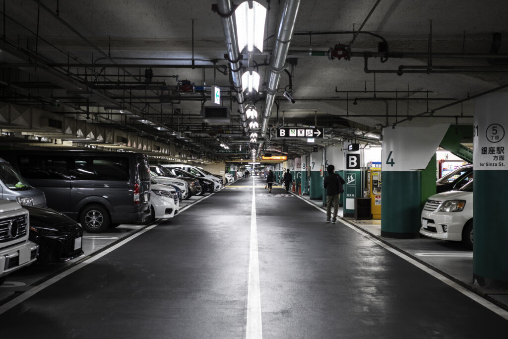 A long, centered view down the driving lane of a dimly lit, underground parking garage with rows of parked cars on both sides. A few people are visible in the distance, emphasizing the need for Parking Lot Surveillance.