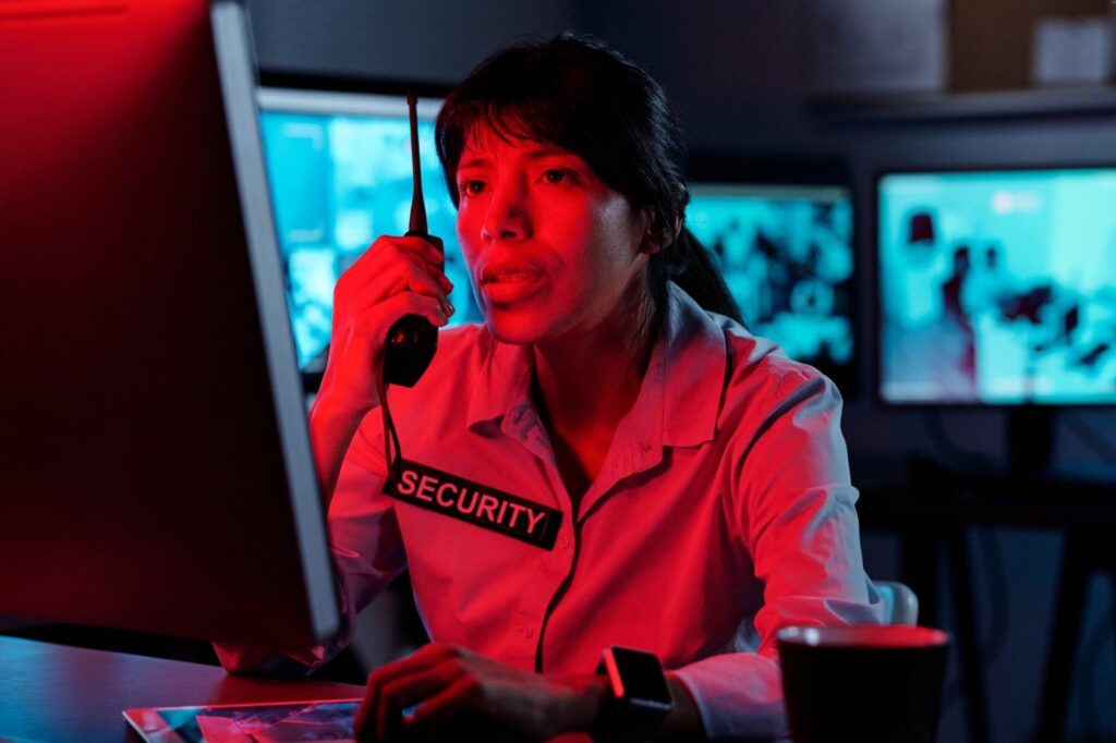 Female security officer speaking into a two-way radio while monitoring video screens, showing active audio surveillance.