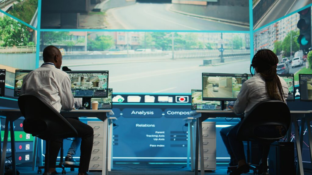 Two security professionals working in a glass-walled office, managing remote CCTV surveillance and analyzing data on multiple screens.