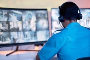 A security guard wearing a blue uniform and headset performing CCTV Monitoring on a large curved multi-view screen.