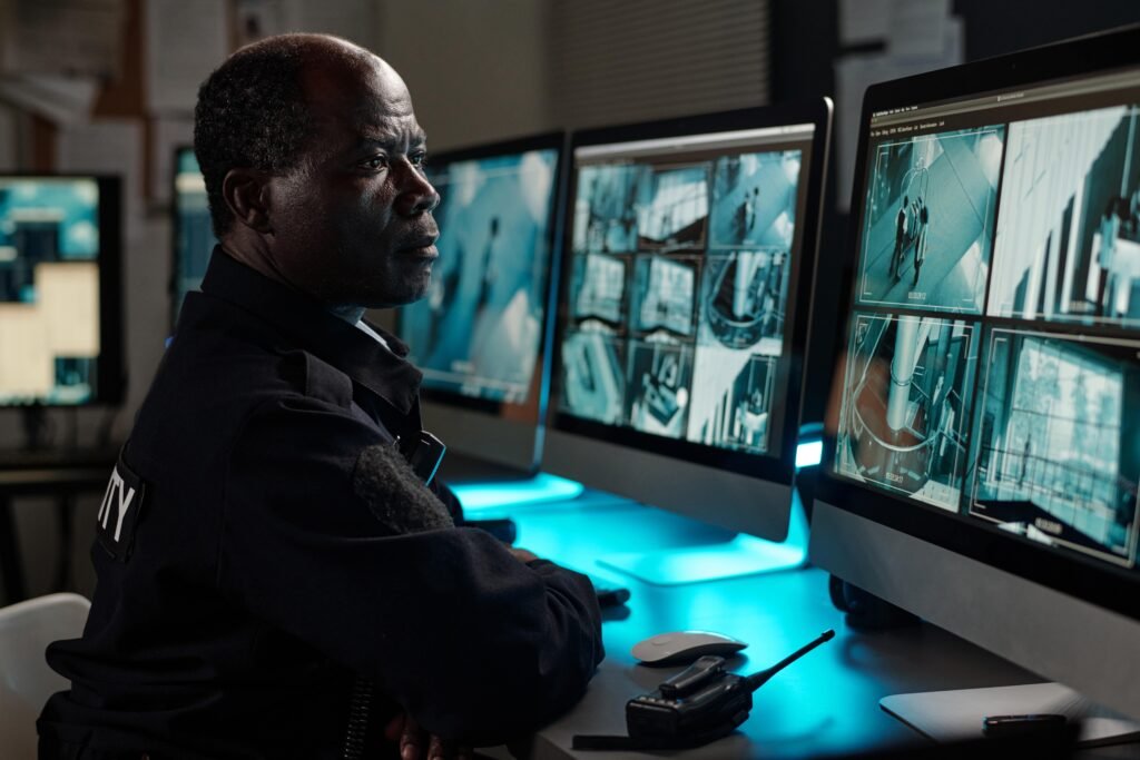 A security professional in a white shirt is shown from behind, monitoring several high-definition screens displaying live CCTV evidence from street and building feeds in a darkened command center