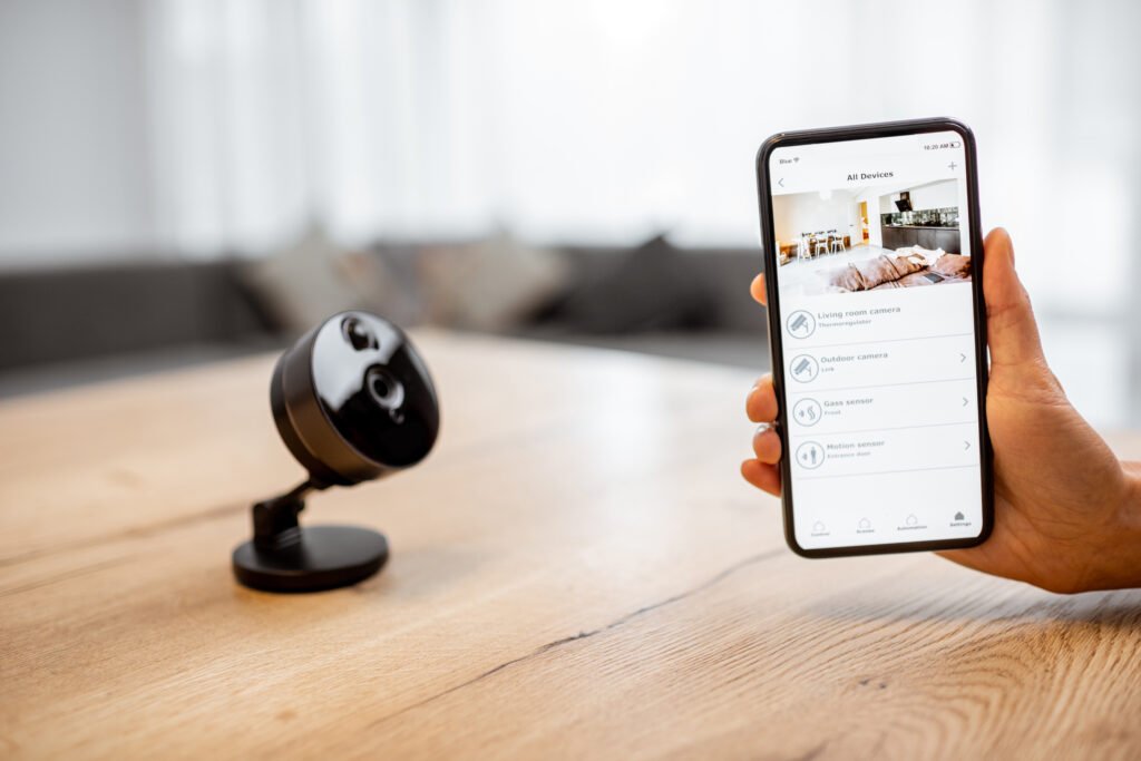 A person holding a smartphone displaying a security monitoring app next to a black indoor camera, illustrating remote access to CCTV surveillance. Security Monitoring Company