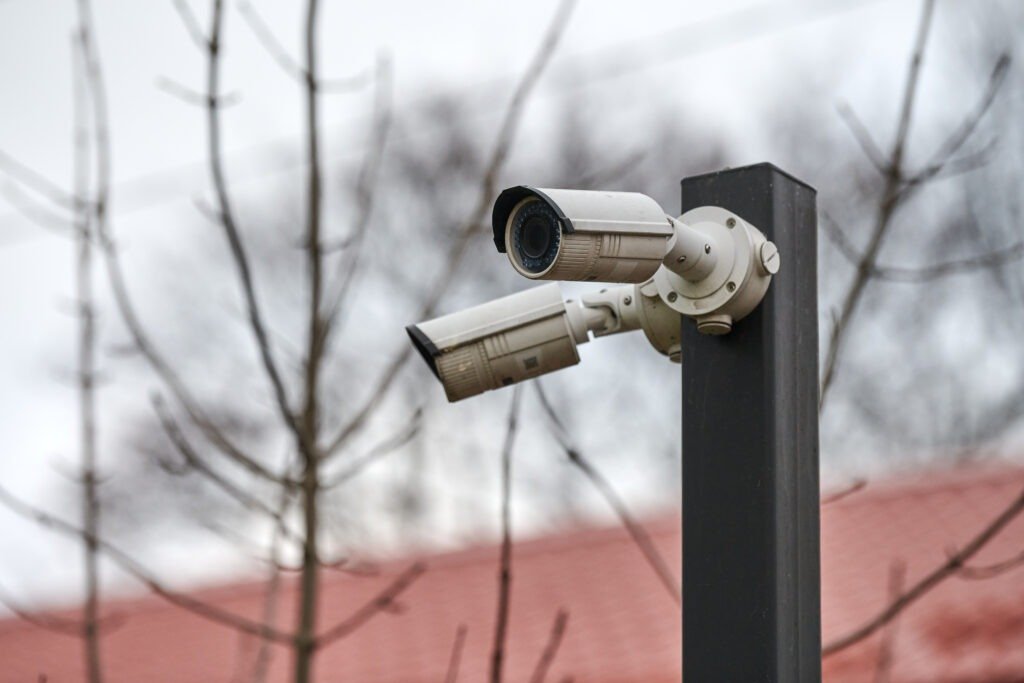 A minimalist isometric graphic of a grey security camera with a yellow light cone on a red background, illustrating Real-Time CCTV Monitoring and Live Surveillance for Property Protection