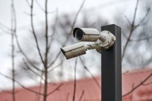 A minimalist isometric graphic of a grey security camera with a yellow light cone on a red background, illustrating Real-Time CCTV Monitoring and Live Surveillance for Property Protection