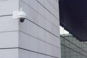 A professional manned security monitoring station illustrating Security Camera Business Benefits, featuring a smiling uniformed guard with a walkie-talkie in front of multiple screens displaying live multi-channel surveillance cameras for business feeds, showing active use of a security camera system for business.