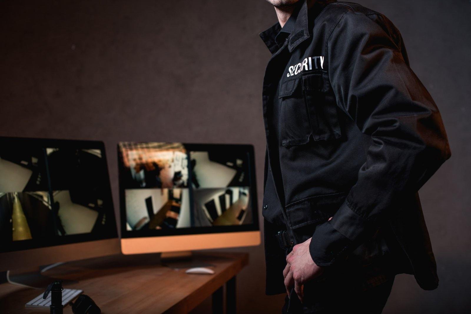 A focused male security operator with a headset in a high-tech control room bathed in blue/magenta light, actively observing a wall of 15 multi-split screens displaying comprehensive CCTV surveillance from diverse commercial video surveillance environments. The scene provides a perfect visual for the debate of CCTV Monitoring vs Security Guards, illustrating how modern technology allows for massive scale in remote security and live security camera monitoring.