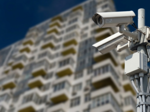 A pair of outdoor bullet security cameras securely mounted on a metal pole in front of a multi-story residential building, highlighting the importance of CCTV monitoring for apartments to ensure comprehensive apartment security and effective building security through a reliable CCTV monitoring service.