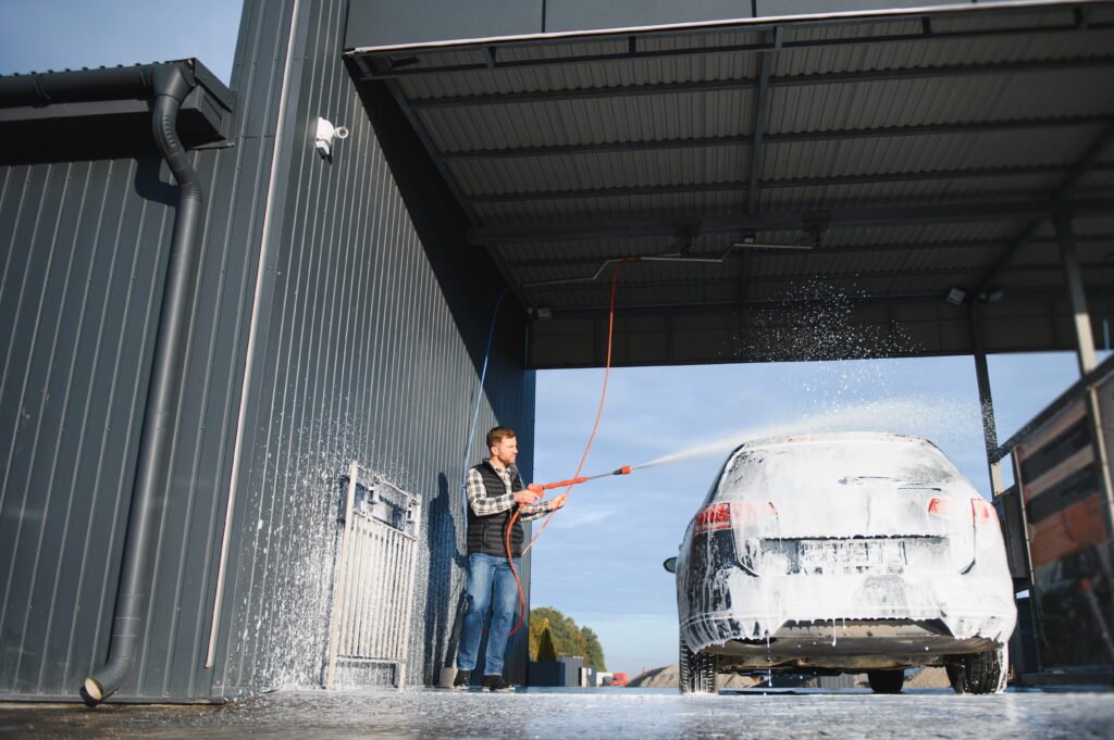 A man uses a high-pressure hose to spray thick foam onto an SUV inside a self-serve bay, while a white bullet camera mounted on the corrugated wall above provides essential CCTV Monitoring for Car Wash operations to ensure continuous carwash security.