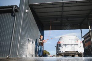 A man uses a high-pressure hose to spray thick foam onto an SUV inside a self-serve bay, while a white bullet camera mounted on the corrugated wall above provides essential CCTV Monitoring for Car Wash operations to ensure continuous carwash security.