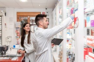 Two retail workers in white lab coats manage shelf inventory behind a point-of-sale counter under the watchful eye of a ceiling-mounted dome camera, highlighting the absolute necessity of CCTV monitoring for dispensaries to maintain strict cannabis security compliance and reliable security surveillance.