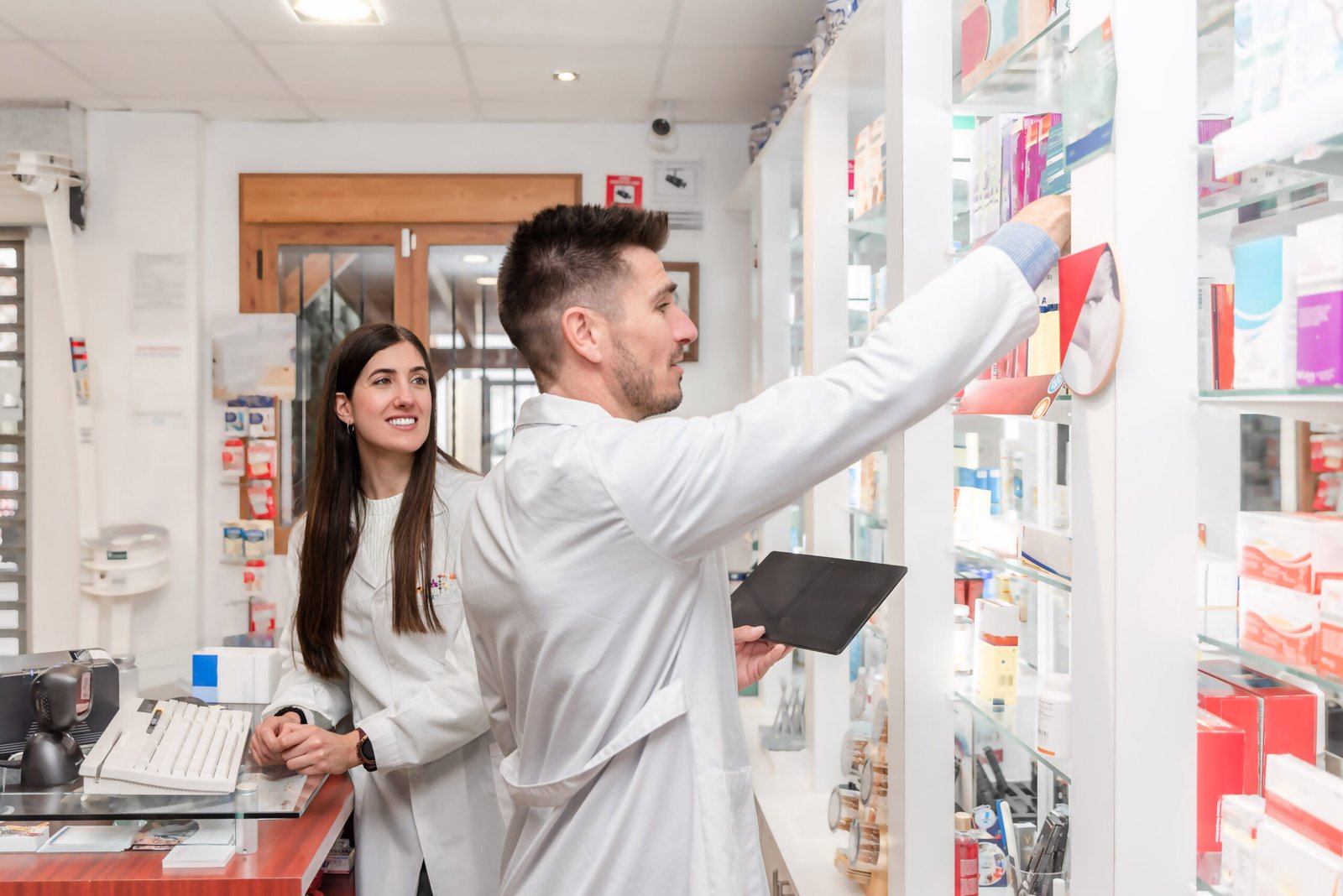 Two retail workers in white lab coats manage shelf inventory behind a point-of-sale counter under the watchful eye of a ceiling-mounted dome camera, highlighting the absolute necessity of CCTV monitoring for dispensaries to maintain strict cannabis security compliance and reliable security surveillance.