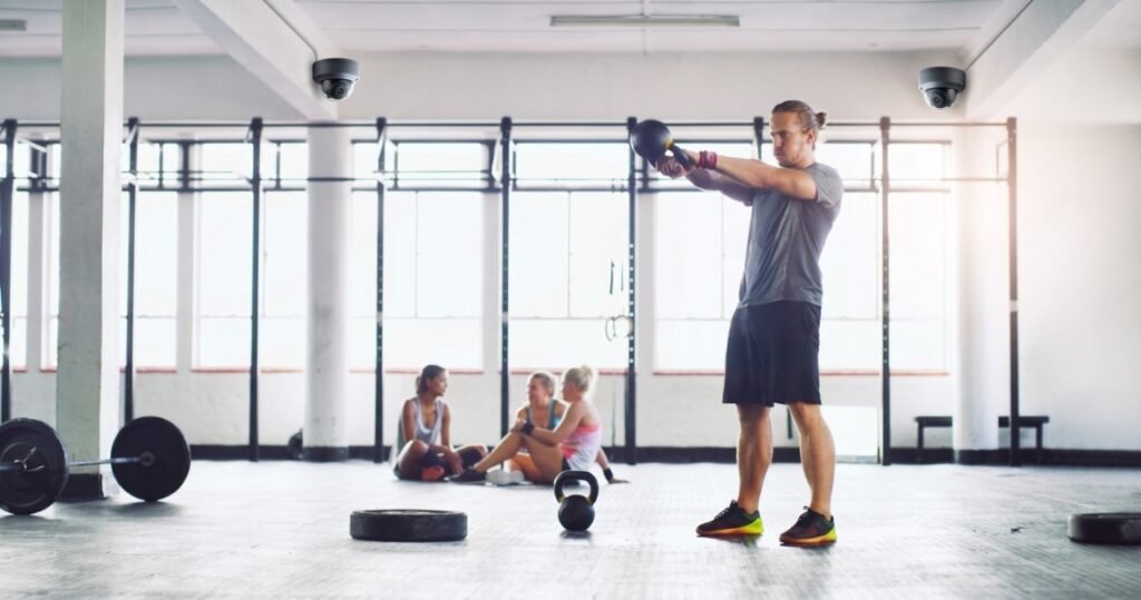 A man actively exercises with a kettlebell on a spacious gym floor while two prominently mounted ceiling dome cameras provide dedicated CCTV Monitoring for Gyms, ensuring member safety through a reliable security service and continuous video surveillance.
