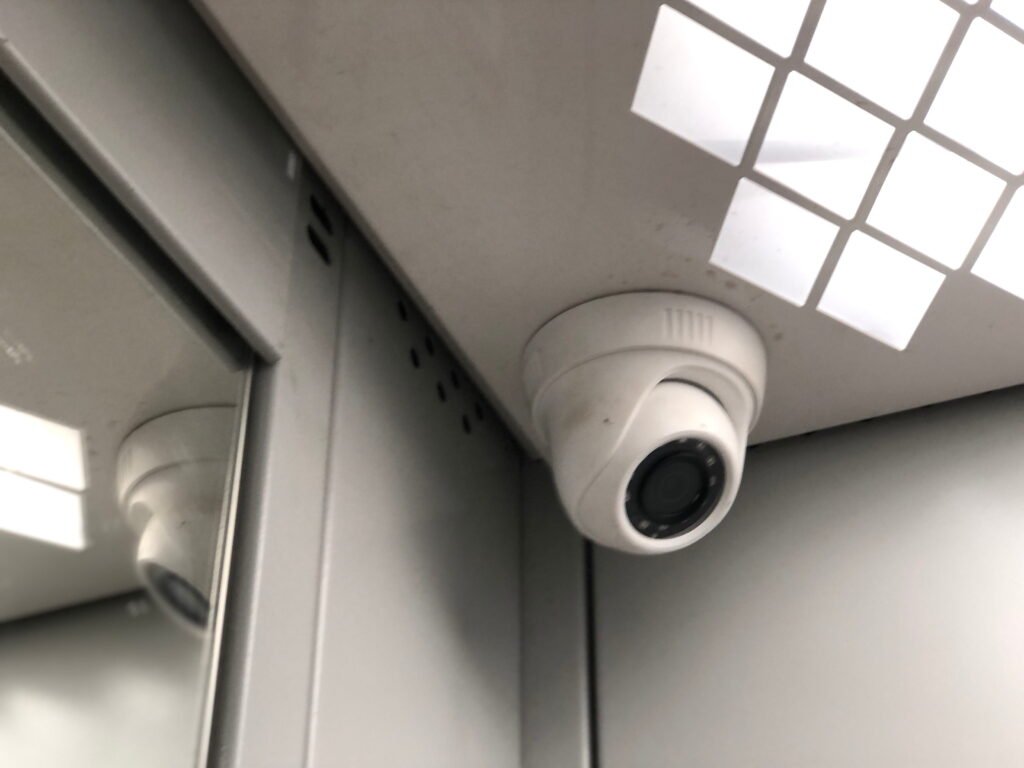 A close-up of a white dome security camera mounted in the corner of a corporate elevator ceiling next to a mirrored wall, demonstrating the strategic placement required for effective CCTV monitoring for office environments by a reliable CCTV monitoring services provider to ensure complete security surveillance.