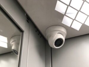 A close-up of a white dome security camera mounted in the corner of a corporate elevator ceiling next to a mirrored wall, demonstrating the strategic placement required for effective CCTV monitoring for office environments by a reliable CCTV monitoring services provider to ensure complete security surveillance.