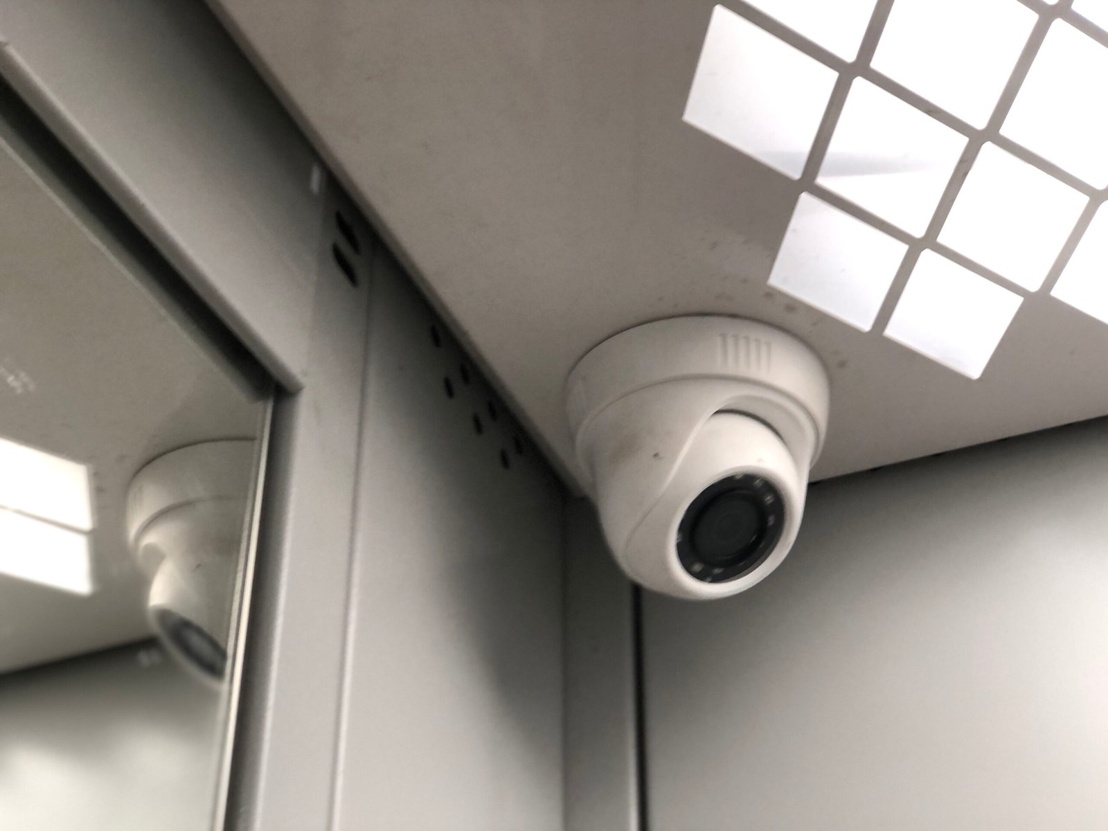 A close-up of a white dome security camera mounted in the corner of a corporate elevator ceiling next to a mirrored wall, demonstrating the strategic placement required for effective CCTV monitoring for office environments by a reliable CCTV monitoring services provider to ensure complete security surveillance.