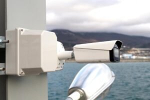 A heavy-duty outdoor security camera mounted on a pole overlooking a body of water, demonstrating the rugged equipment needed for effective CCTV monitoring for ship docks to protect valuable assets, much like the wealth protection advice found in our Sell Gold Coins Guide.