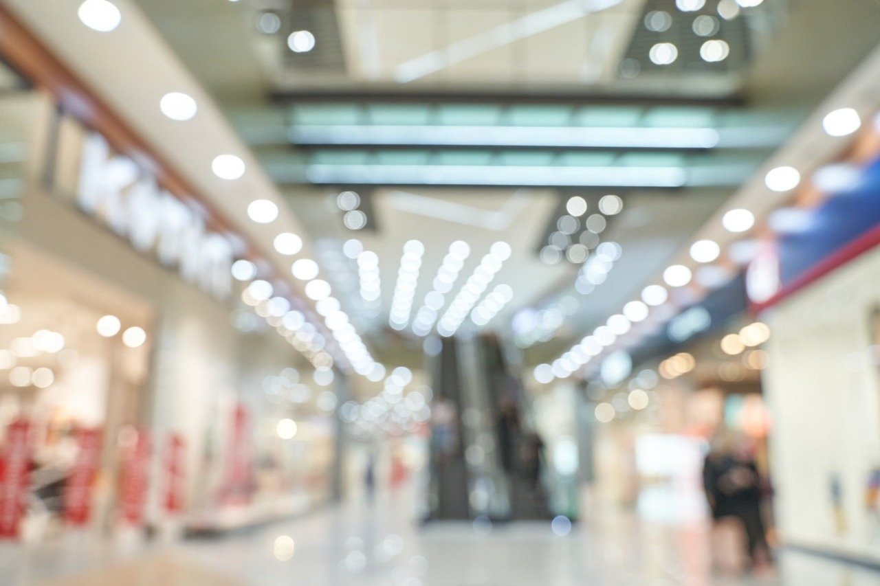 A blurred background view of a brightly lit interior of a shopping center with escalators, representing the expansive public areas that require comprehensive CCTV Monitoring for Shopping Malls using a reliable cctv monitoring service and commercial security cameras to ensure optimal shopping center security.
