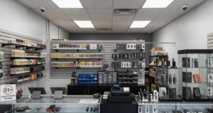 Alt Text: An interior view of a brightly lit retail store with glass display cases and shelves fully stocked with e-liquids, featuring two ceiling-mounted dome cameras that demonstrate the vital need for reliable CCTV monitoring for vape shops using remote CCTV monitoring and expert surveillance services.