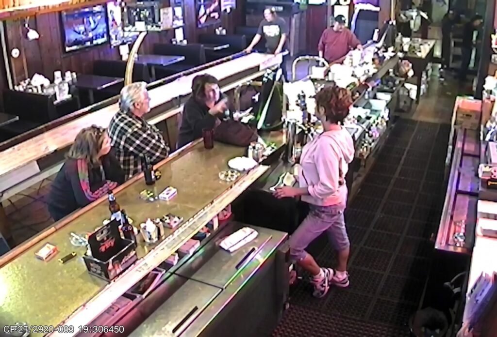 A high-angle security camera view overlooking a bartender and seated patrons inside a neighborhood pub, illustrating the practical necessity of CCTV monitoring for bars to provide reliable security surveillance and dedicated surveillance services for a safe nightlife environment.