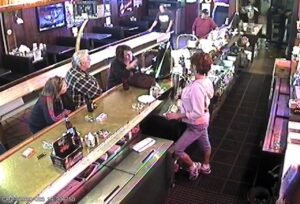 A high-angle security camera view overlooking a bartender and seated patrons inside a neighborhood pub, illustrating the practical necessity of CCTV monitoring for bars to provide reliable security surveillance and dedicated surveillance services for a safe nightlife environment.