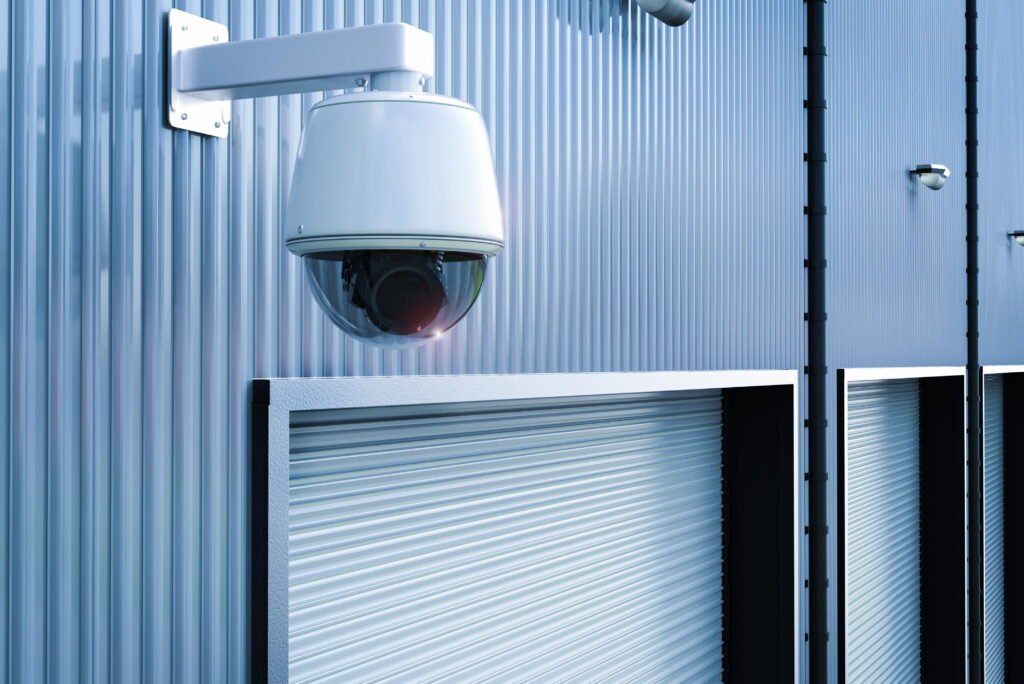 A close-up of a prominent white dome security camera mounted on a corrugated metal wall directly above a row of closed roll-up doors, demonstrating the vital role of CCTV monitoring for storage to ensure comprehensive storage facility security and continuous surveillance monitoring.