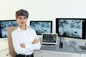 A professional female security specialist with arms crossed sitting in front of multiple monitors displaying split-screen multi-camera feeds, illustrating the operational setup of advanced business cctv systems.