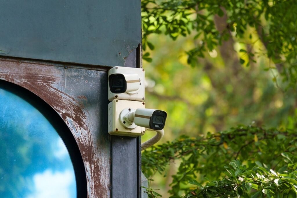 A close-up shot of two white outdoor bullet surveillance cameras mounted vertically on a rustic, weathered metal beam next to a circular reflective blue window with vibrant green foliage in the background, illustrating why overlooking hidden operational overheads can cause Your System Costs to skyrocket beyond initial hardware estimates.
