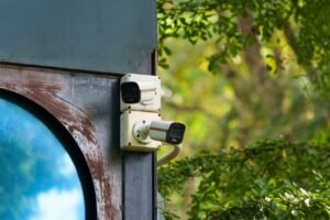 A close-up shot of two white outdoor bullet surveillance cameras mounted vertically on a rustic, weathered metal beam next to a circular reflective blue window with vibrant green foliage in the background, illustrating why overlooking hidden operational overheads can cause Your System Costs to skyrocket beyond initial hardware estimates.
