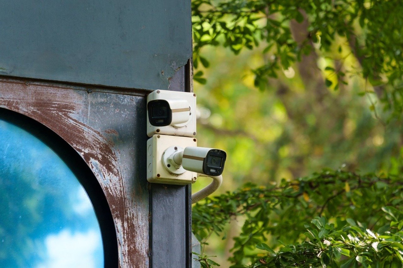 A close-up shot of two white outdoor bullet surveillance cameras mounted vertically on a rustic, weathered metal beam next to a circular reflective blue window with vibrant green foliage in the background, illustrating why overlooking hidden operational overheads can cause Your System Costs to skyrocket beyond initial hardware estimates.