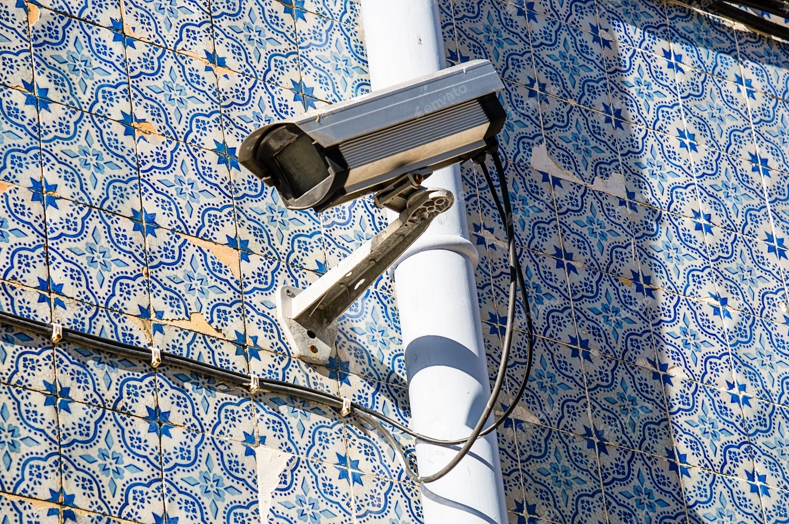 An outdoor industrial box security camera mounted on a white support pole against a highly detailed blue and white patterned ceramic tile wall, illustrating the physical setup of commercial cctv systems where a hidden cctv footage mistake can severely impact an organization's legal protection.