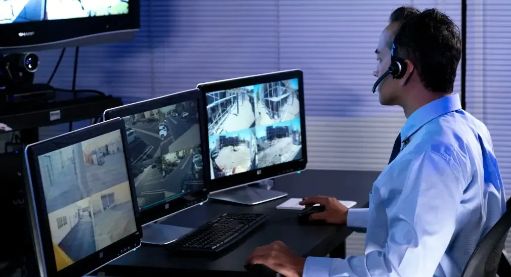 Security technician wearing a headset, monitoring live Remote CCTV Monitoring footage on multiple computer screens in a control room.
