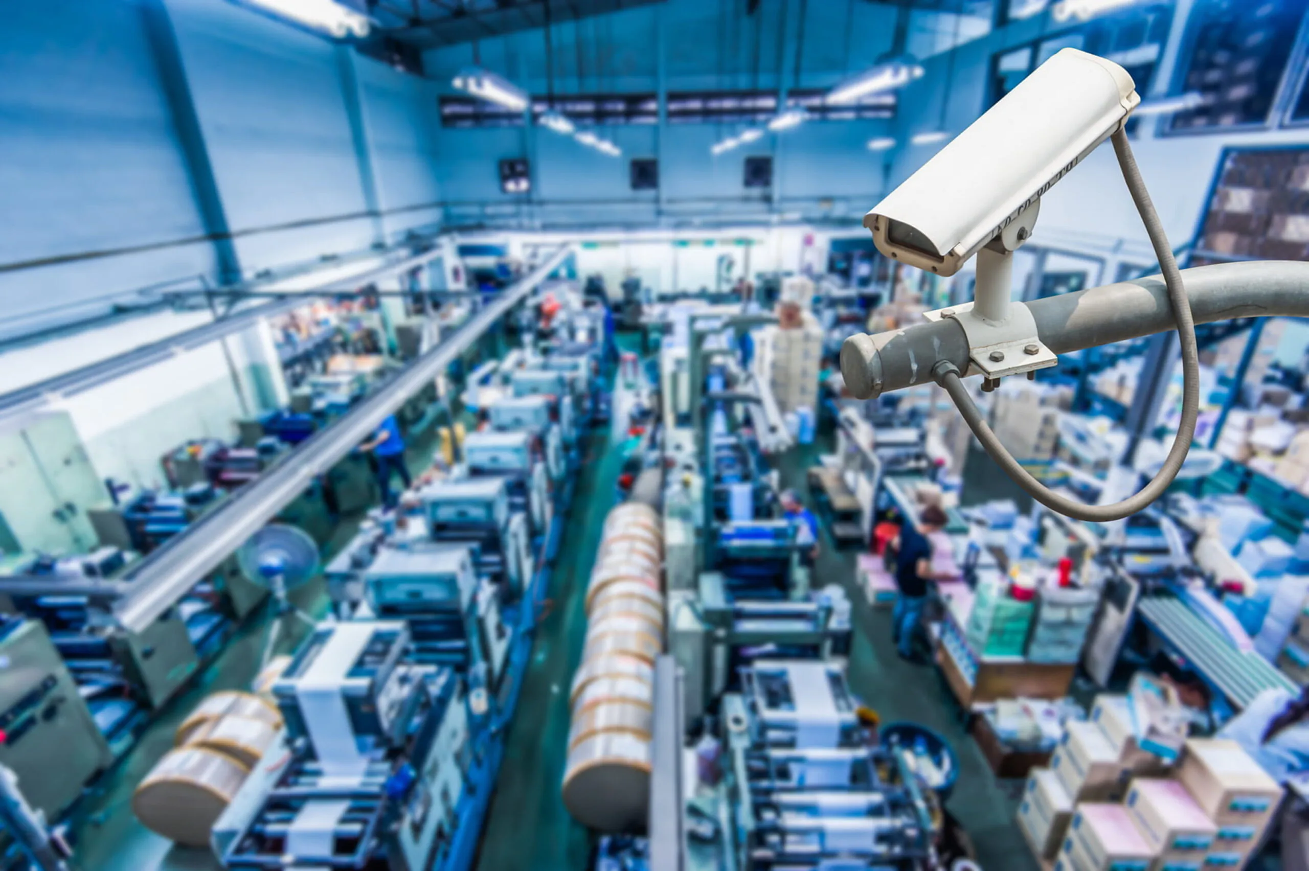 A white outdoor CCTV camera oversees a bustling, brightly lit industrial or warehouse facility with various machinery, emphasizing constant surveillance for industrial and warehouse security.