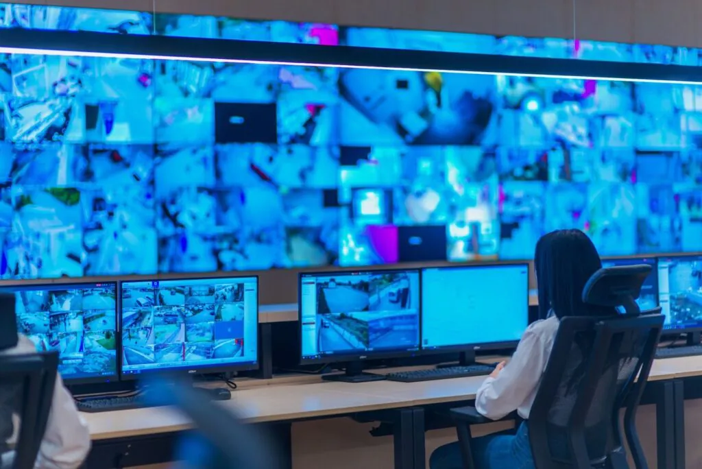 A security operator with dark hair, viewed from behind, sits at a desk in a control room, and monitor villas, intently monitoring multiple large screens displaying various live CCTV feeds with a blue hue.