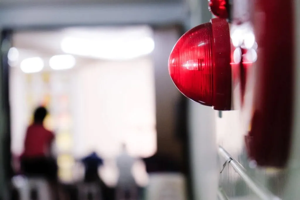 A bright red emergency alarm light is in sharp focus on the right, mounted on a wall, with a blurred background showing people in a brightly lit indoor space.